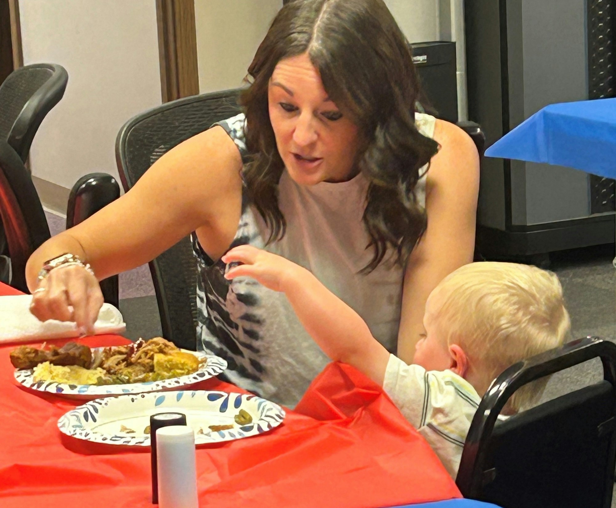 A woman serving food with a toddler watching at a table with a red tablecloth.