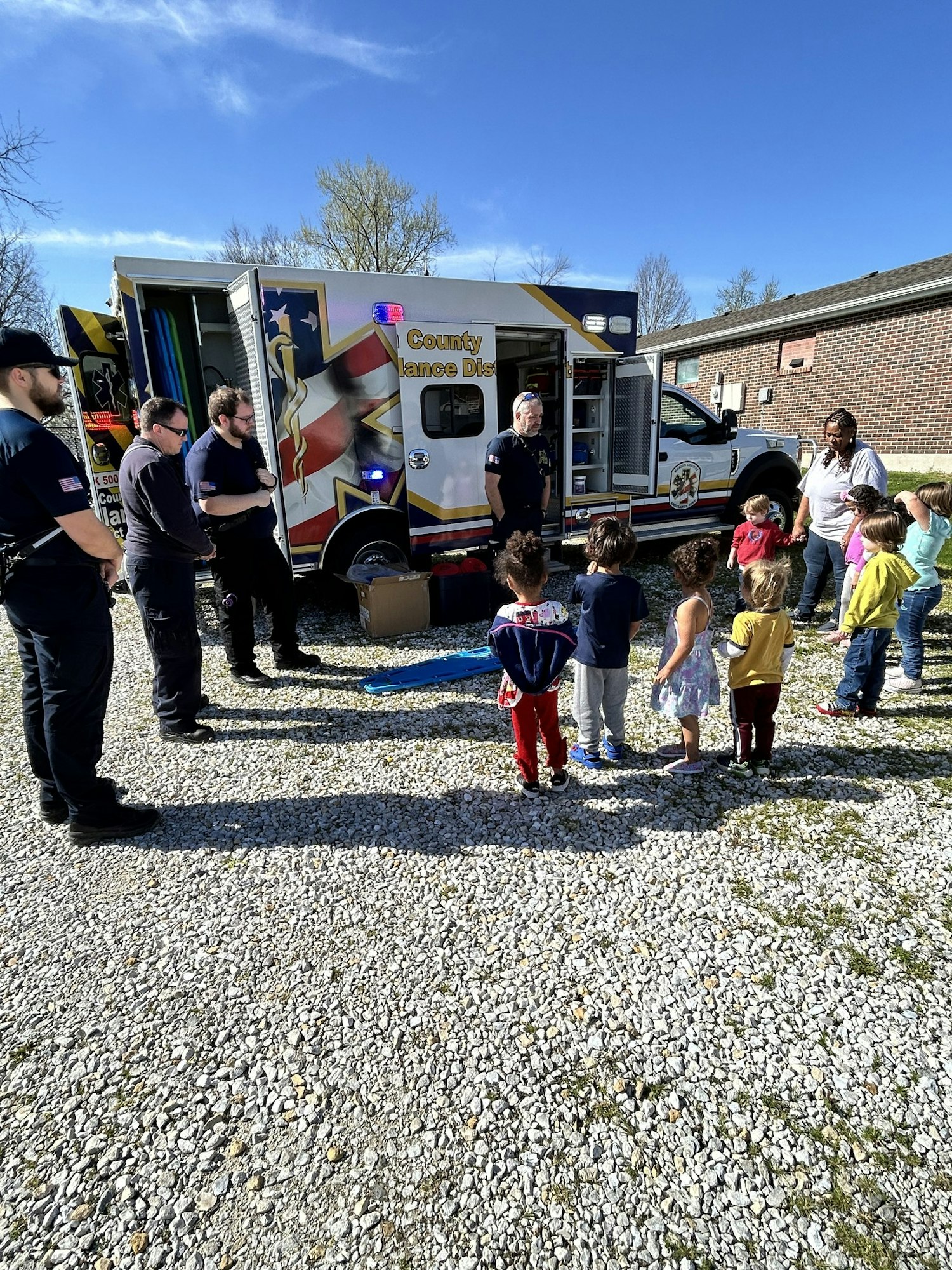 An ambulance with open doors and emergency responders teaching a group of children outside.