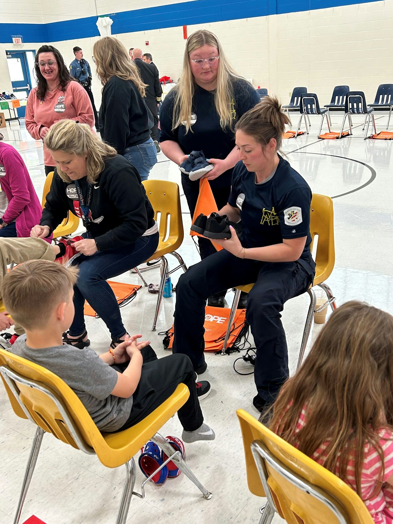 Adults fitting children with shoes in a gym-like setting, with onlookers and emergency service uniforms visible.