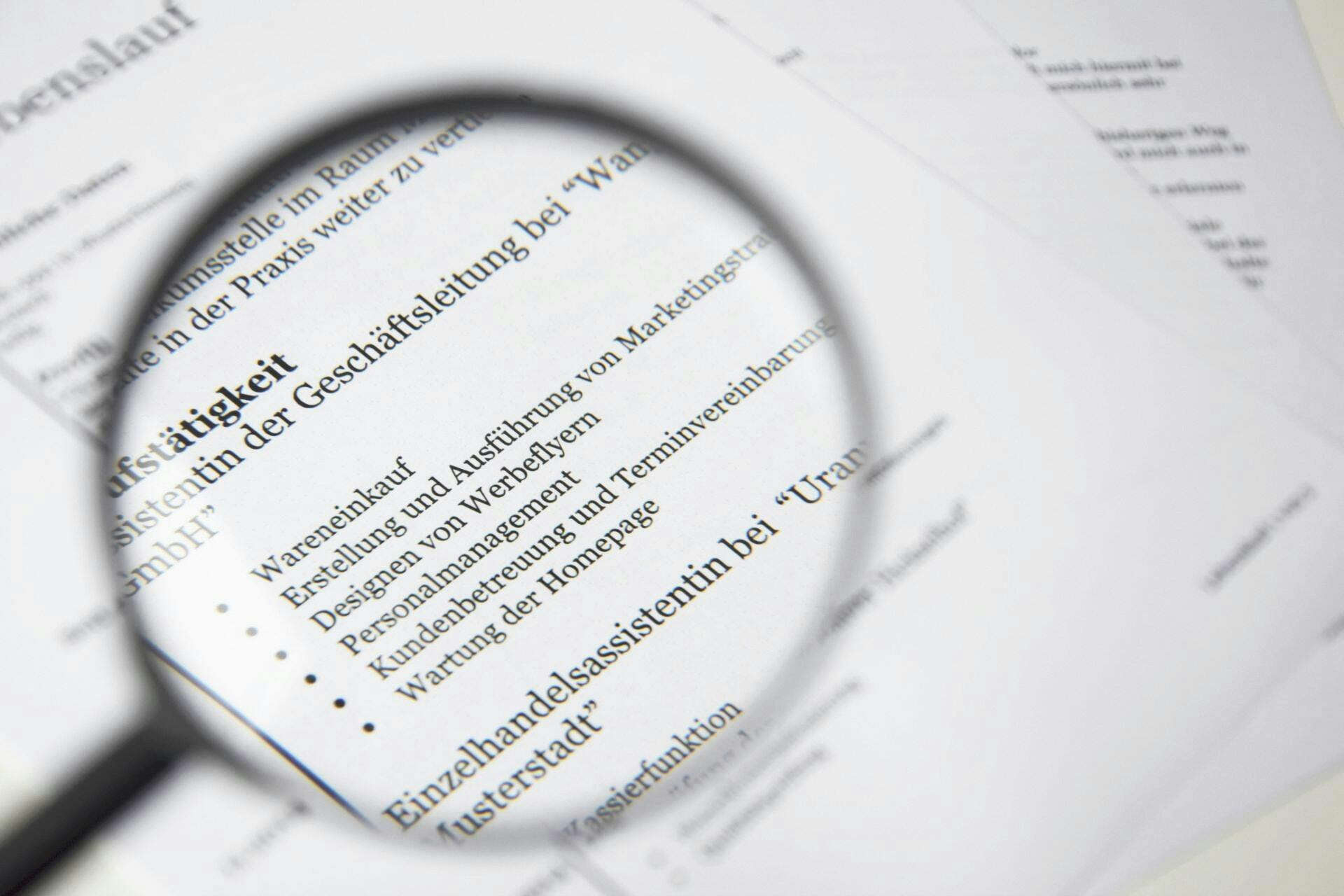 The image shows a magnifying glass highlighting text on printed documents, likely a resume or job description in German.