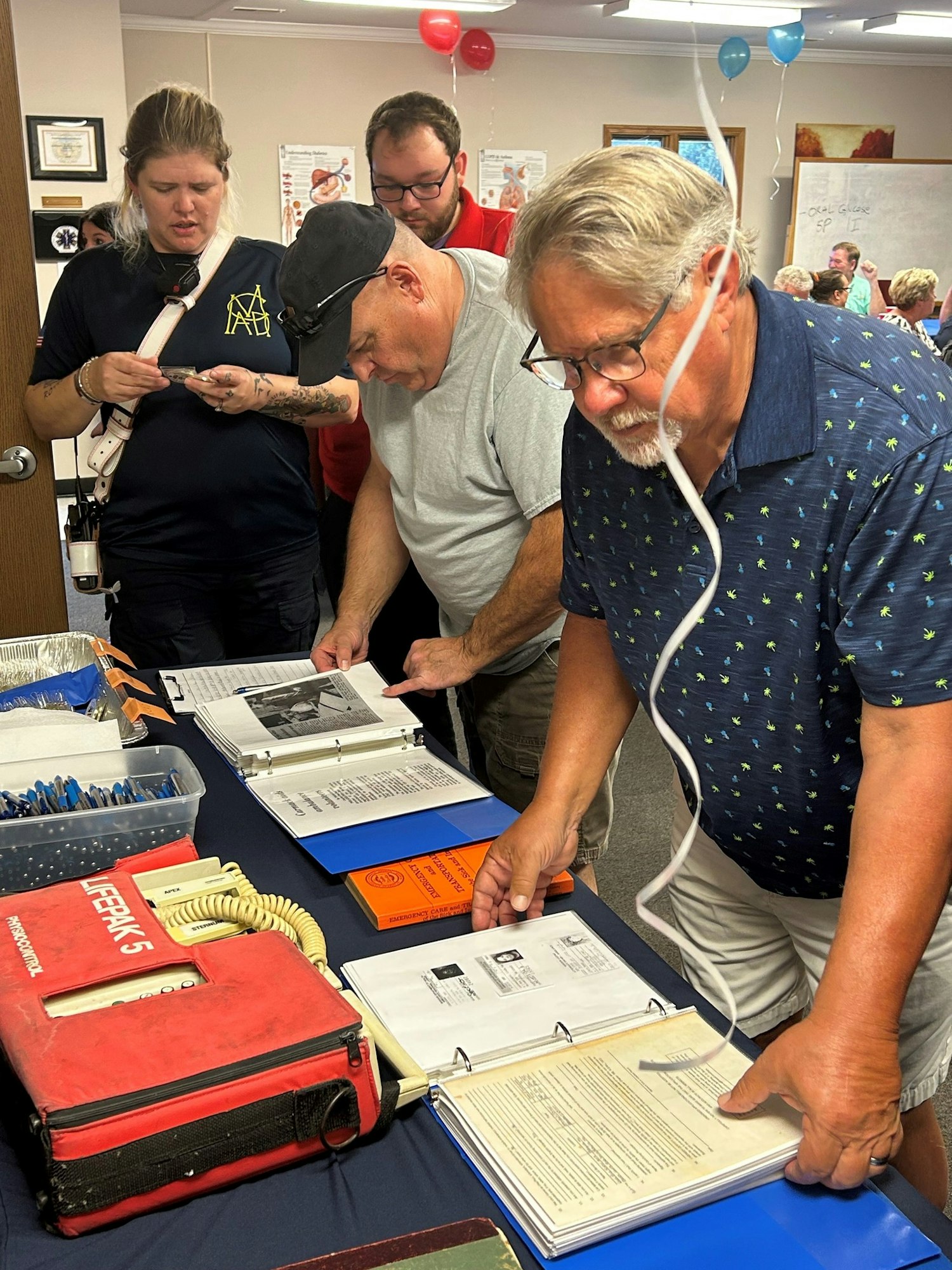 People examining documents and binders on a table at a gathering with balloons in the background.