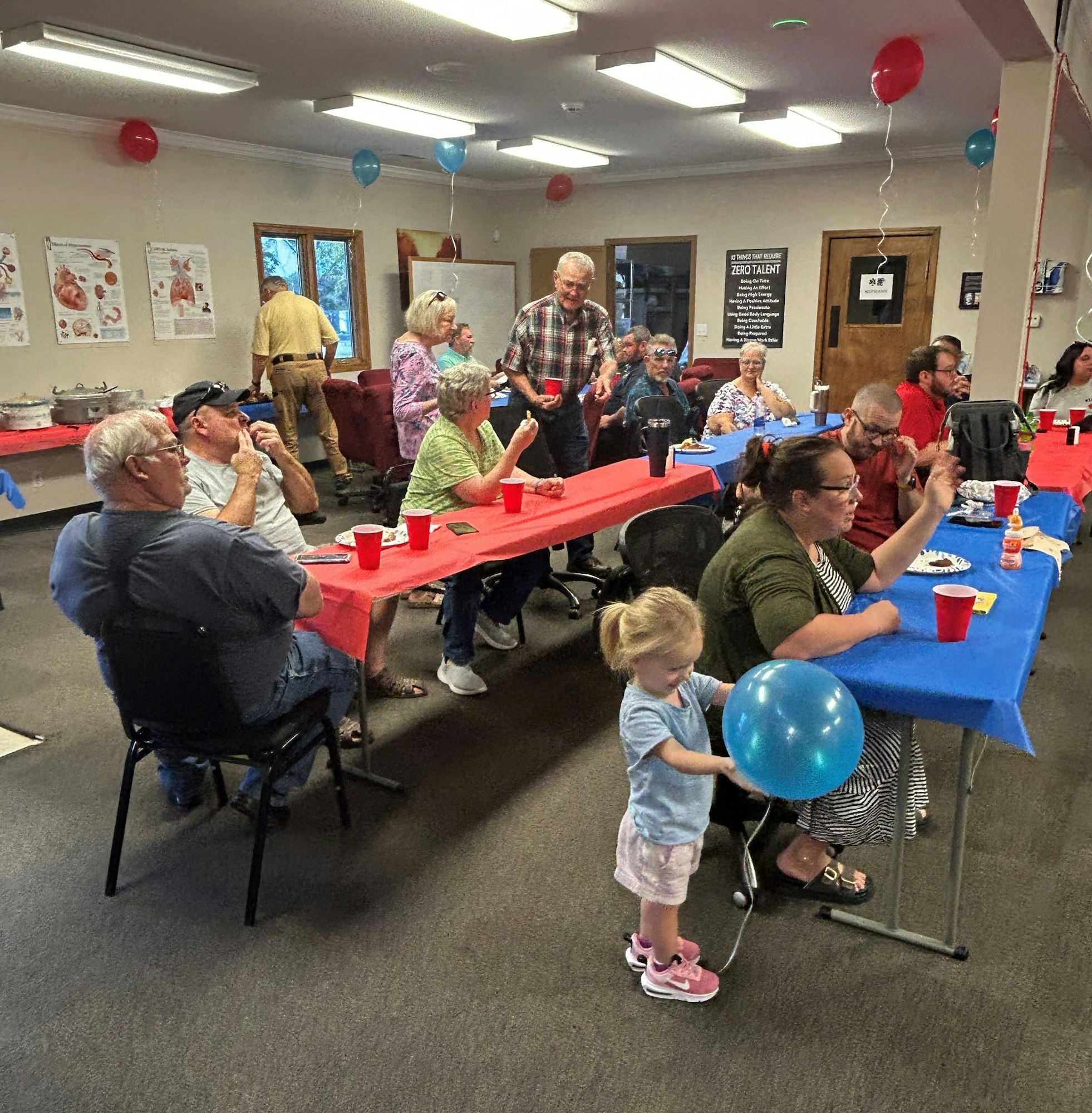 A gathering with people sitting at tables with red cups, decorations, and a child holding a balloon.