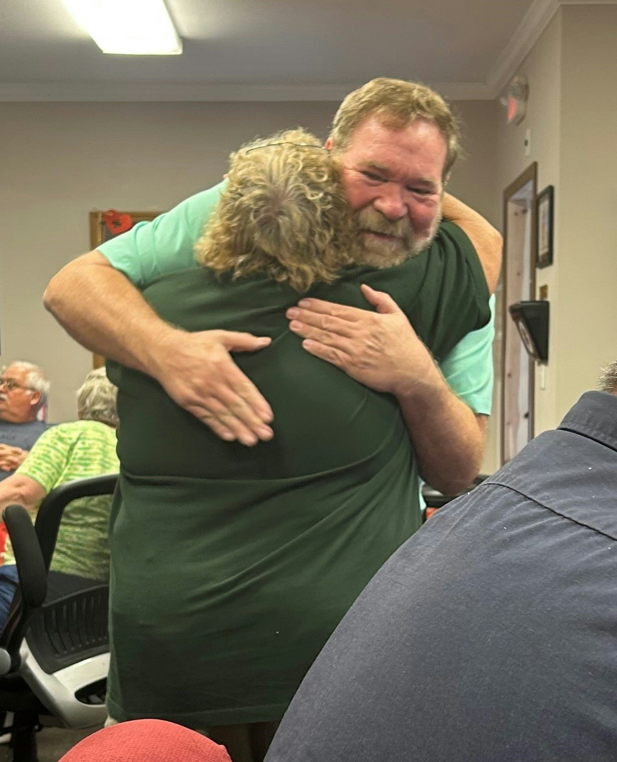 Two people are hugging each other warmly in a room with seated individuals in the background.