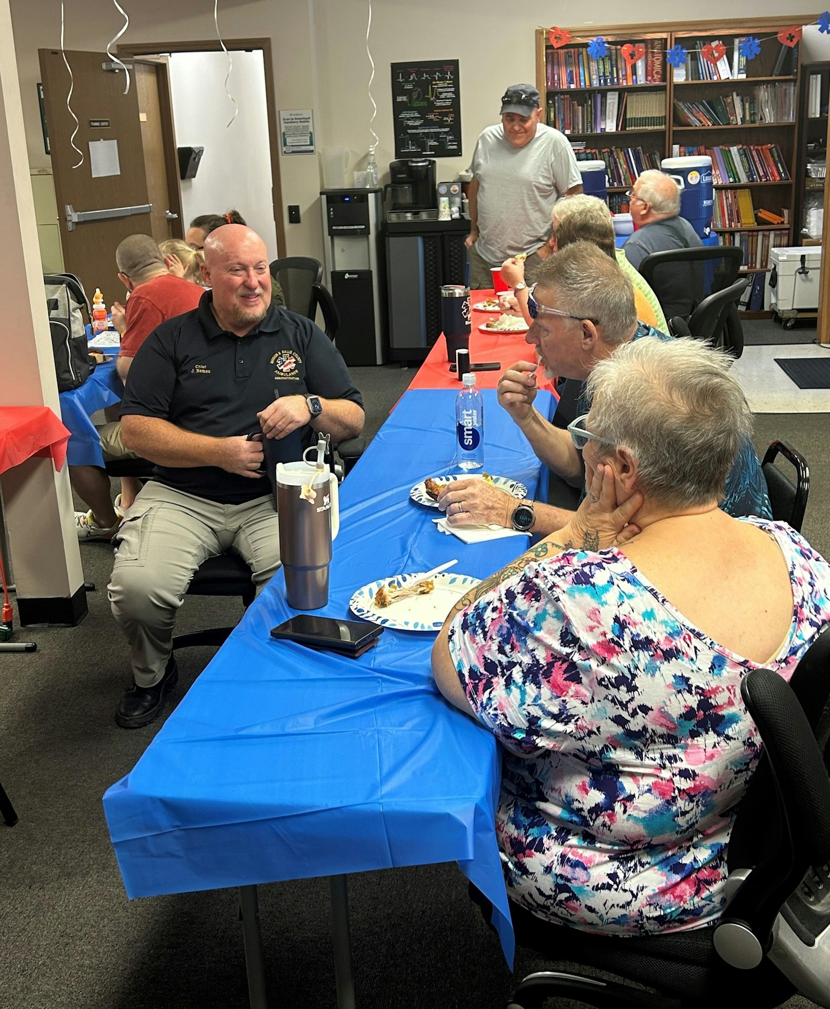People gathered at tables with blue covers, enjoying a meal and conversation in a room with decorations and bookshelves.
