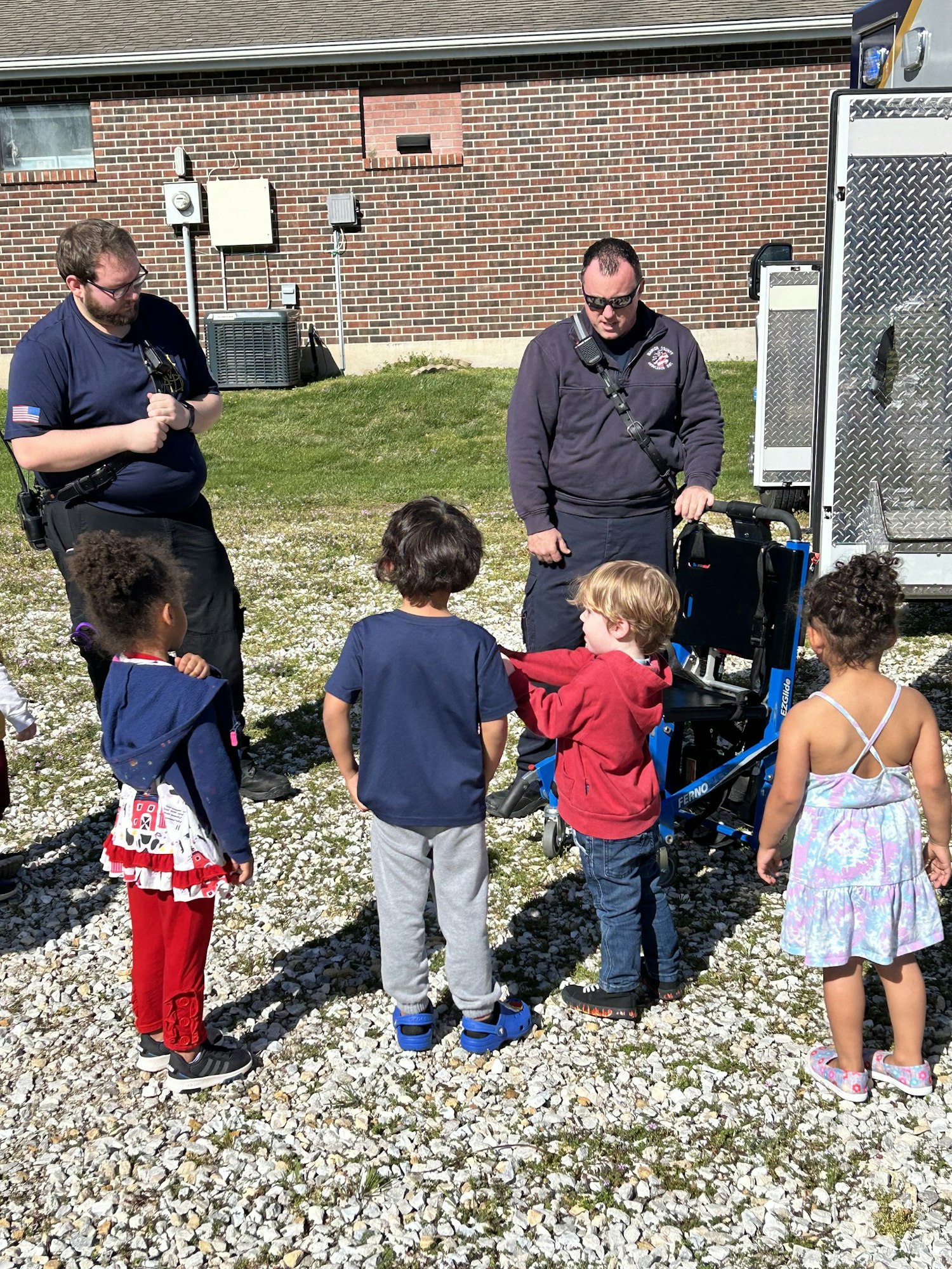 Two adults, likely paramedics, are showing equipment to a group of curious children outdoors.