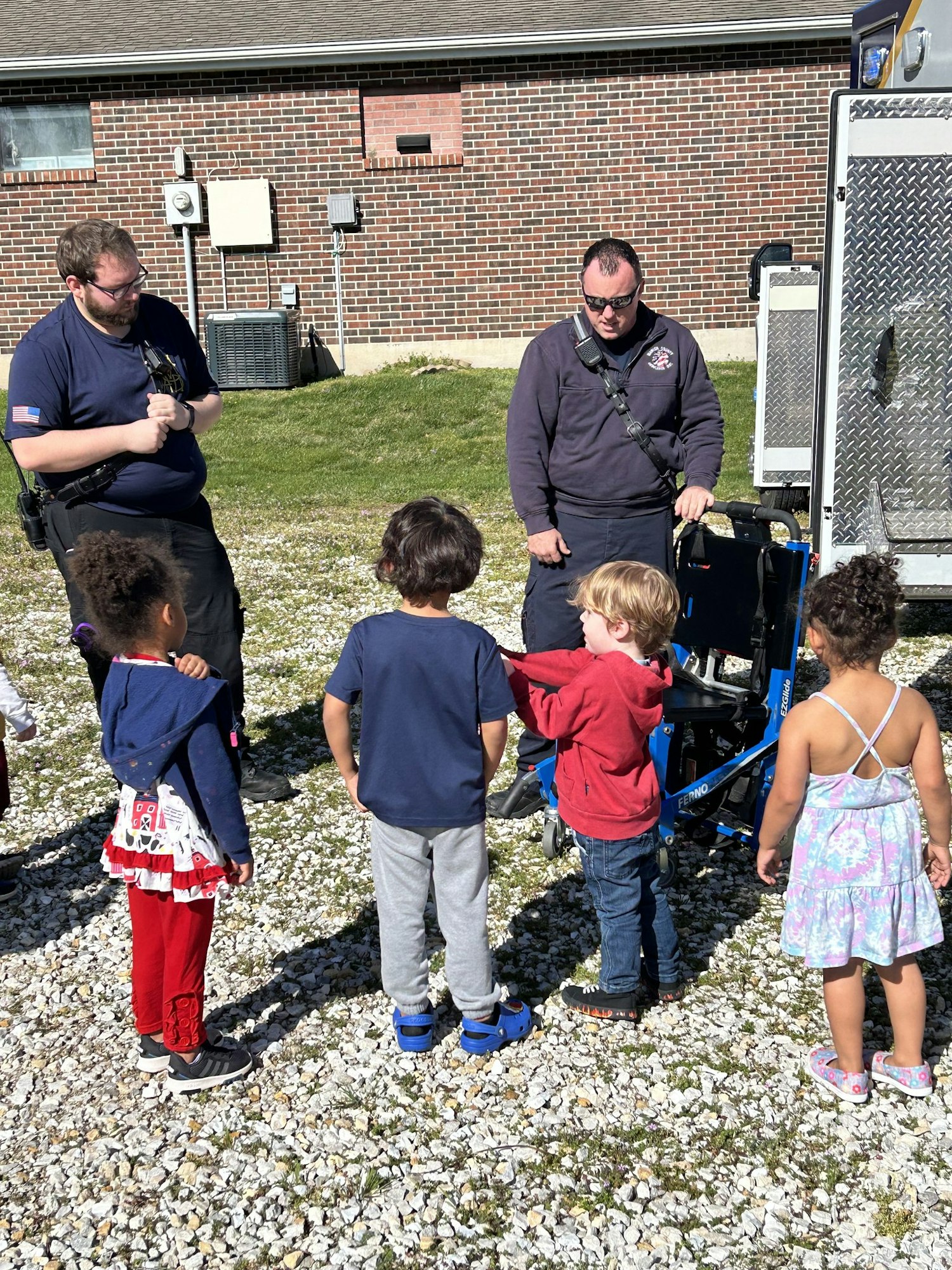 Two adults, likely paramedics, are showing equipment to a group of curious children outdoors.