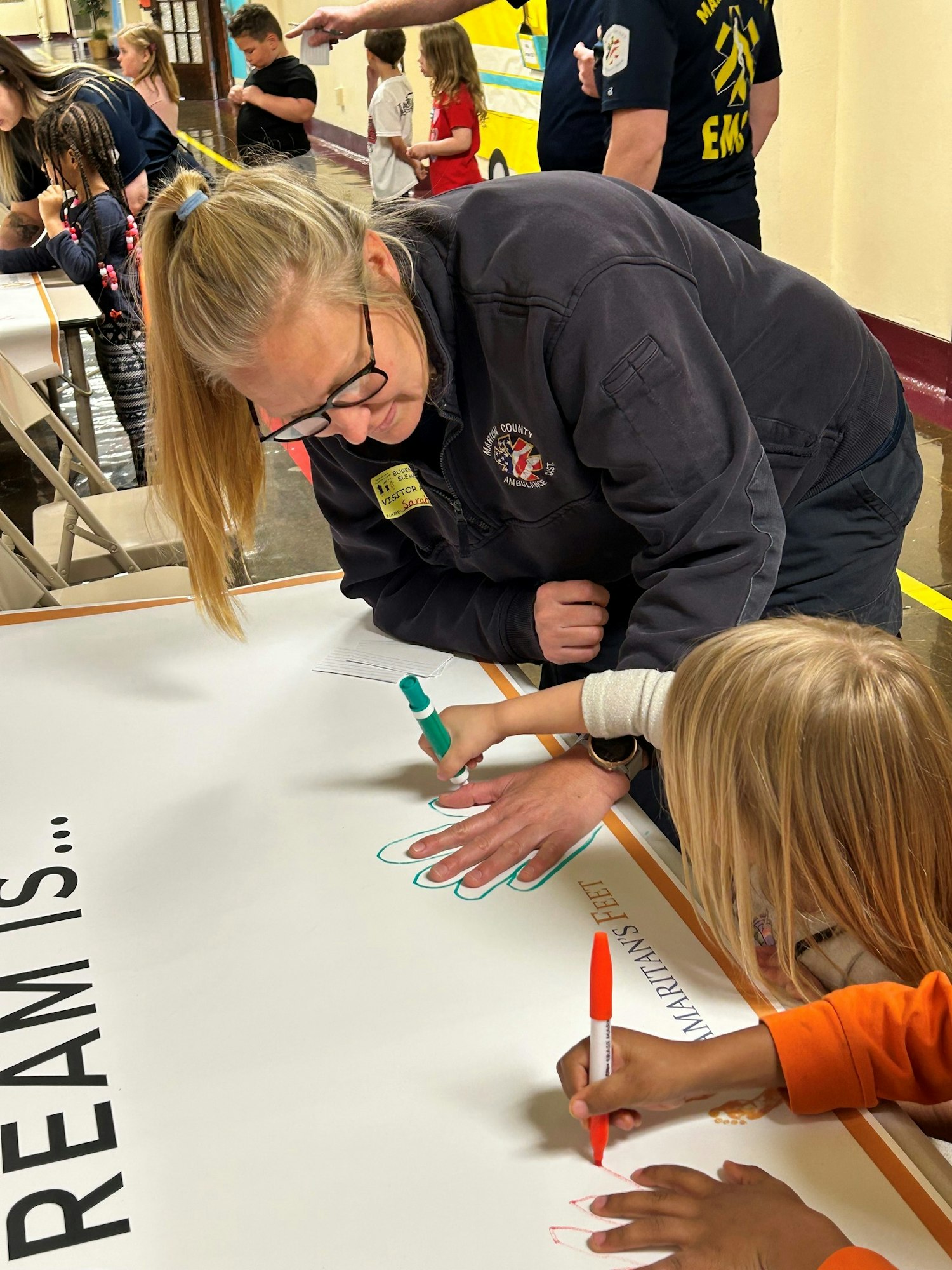 Adult and child tracing hands on a poster with text "DREAMS IS..." at a community event.