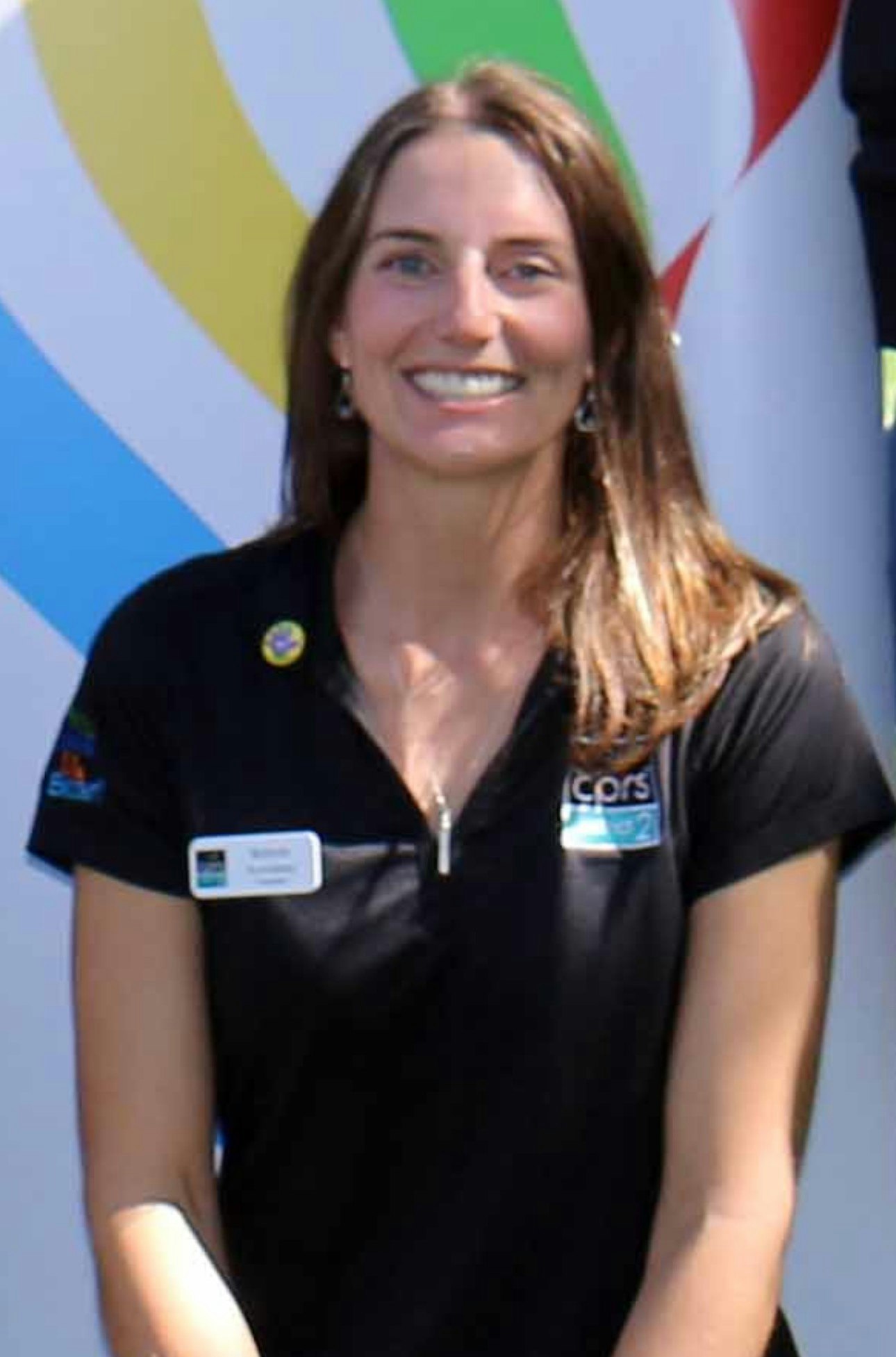 A smiling woman in a black polo shirt with a name tag and logo, against a colorful backdrop.