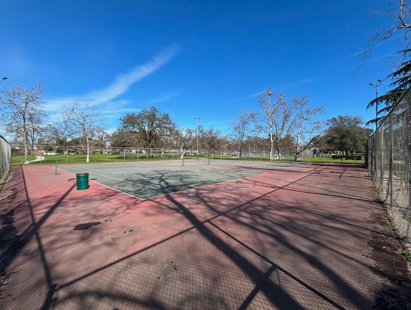 An empty tennis court surrounded by trees and fencing under a clear blue sky.