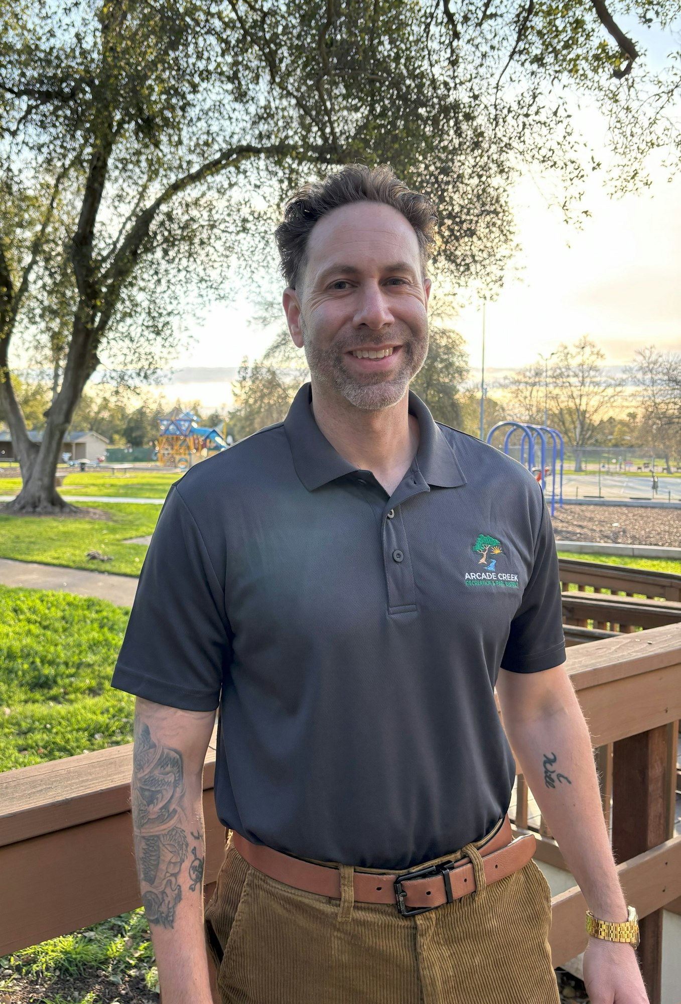A person with tattoos smiles in front of a park background, wearing a polo shirt with "Arcade Creek" logo.