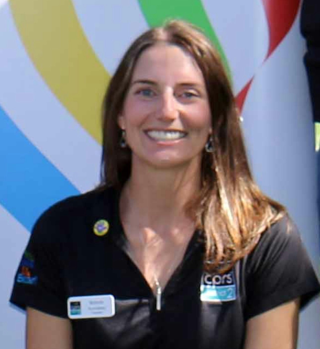 A smiling woman in a black shirt with badges, standing in front of a colorful background.