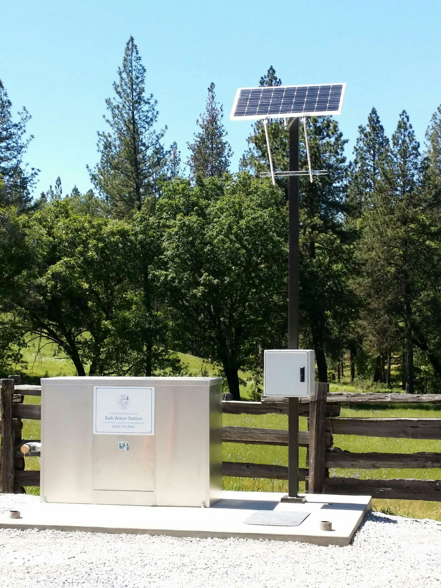 A small solar-powered utility station with a control panel and a pole-mounted solar panel stands on a concrete slab next to a wooden fence. The background features green trees and a clear blue sky, suggesting a rural or forested setting.