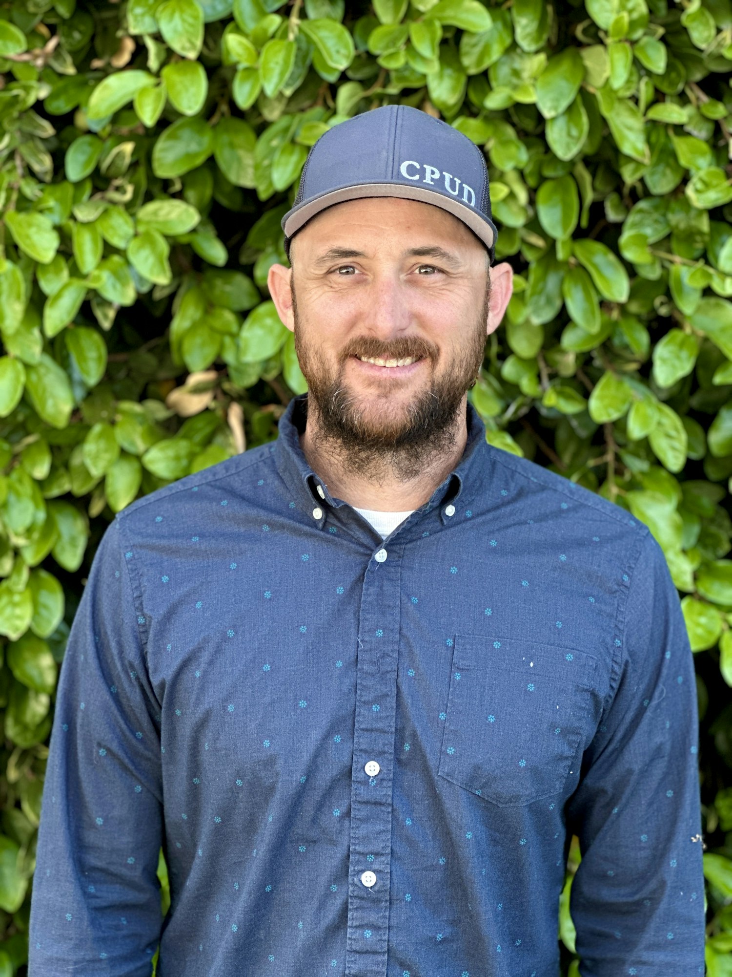 A person in a blue button-up shirt and cap stands in front of a leafy background.