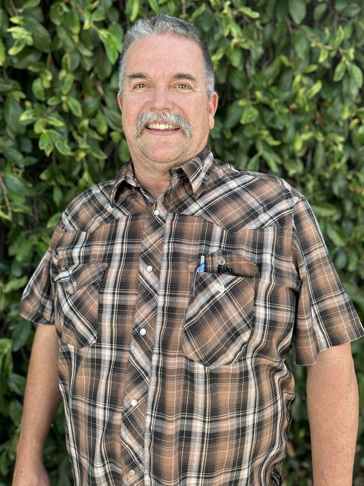 A smiling man with a mustache wearing a plaid shirt against a green leafy background.