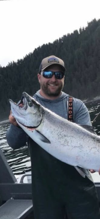 A man on a boat proudly holds a large fish, surrounded by water and forested hills in the background.