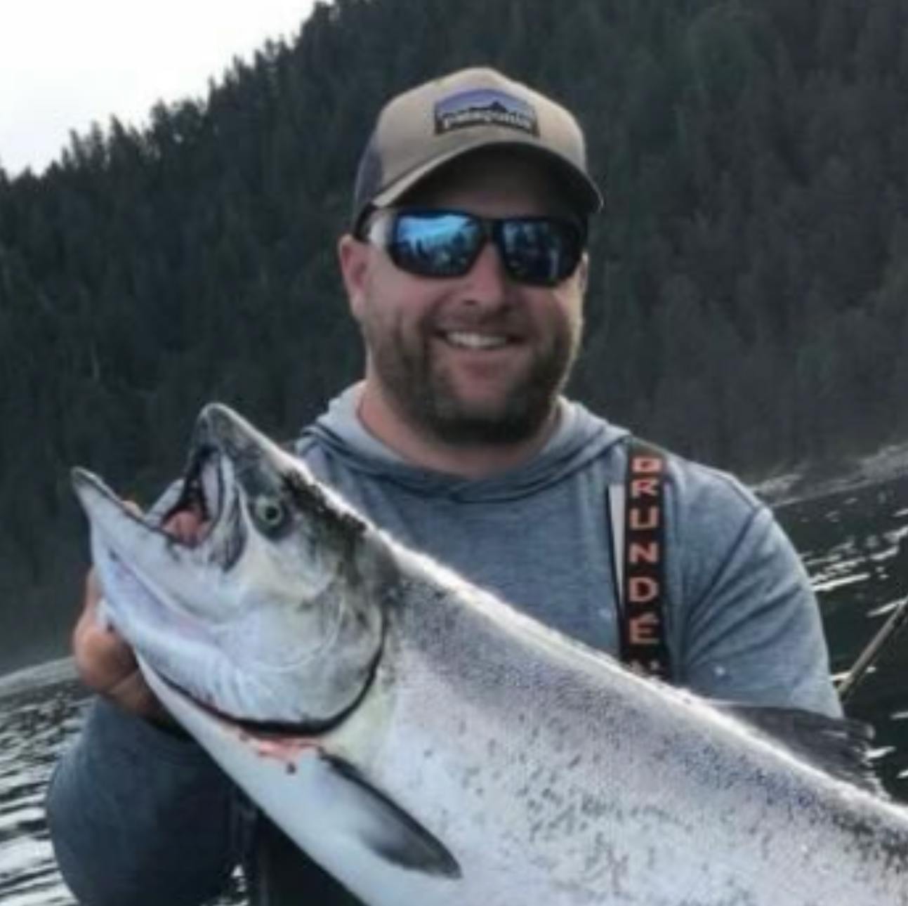 A man on a boat proudly holds a large fish, surrounded by water and forested hills in the background.