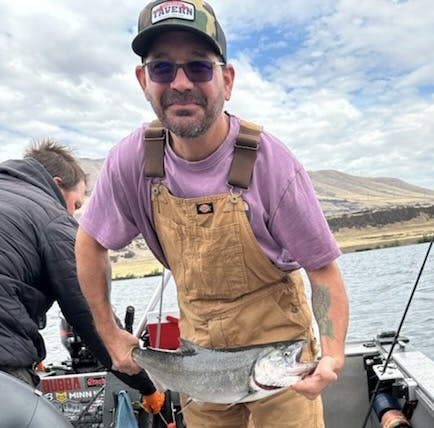 A person on a boat holding a fish, wearing overalls and a cap, with another person nearby and water in the background.