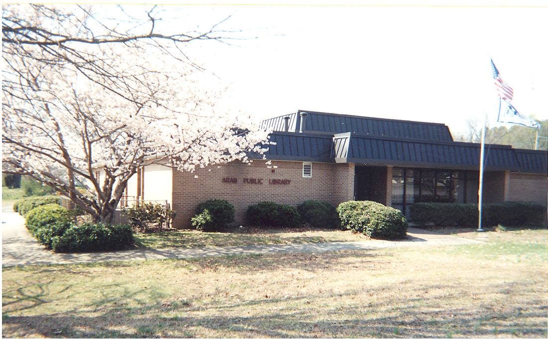 A library building with a flagpole, surrounded by trees and bushes under a clear sky.