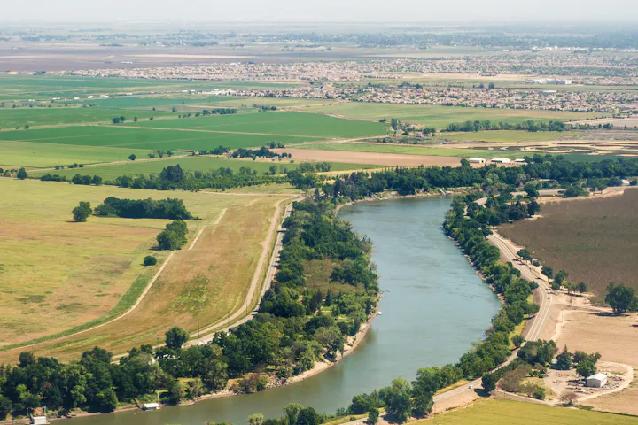An aerial view showing a winding river surrounded by lush green fields, roads, and scattered houses in the background.