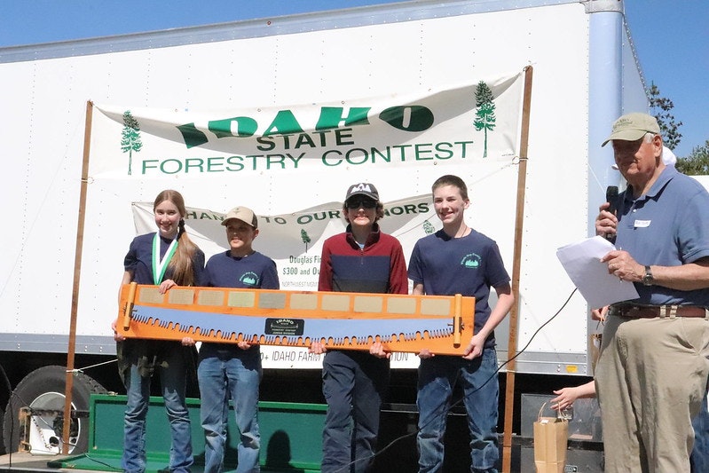 Group of people posing with a saw in front of an "Idaho State Forestry Contest" banner, with a man speaking on a microphone.