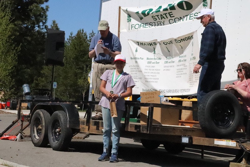 A person stands with a medal and trophy on a stage at an outdoor forestry contest event.