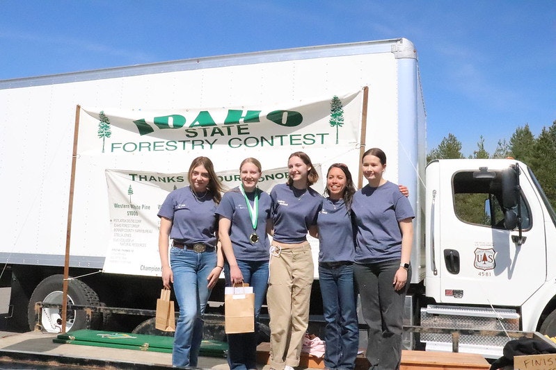 Five people in matching shirts stand together in front of an "Idaho State Forestry Contest" banner, with a truck in the background.