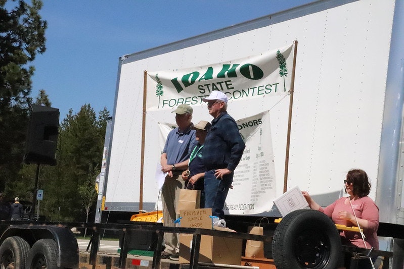People on a platform at the Idaho State Forest Contest, with a sign and awards displayed around them.