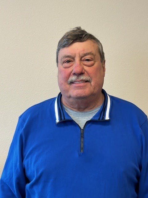 A man in a blue zip-up with a collar, smiling, against a plain background.
