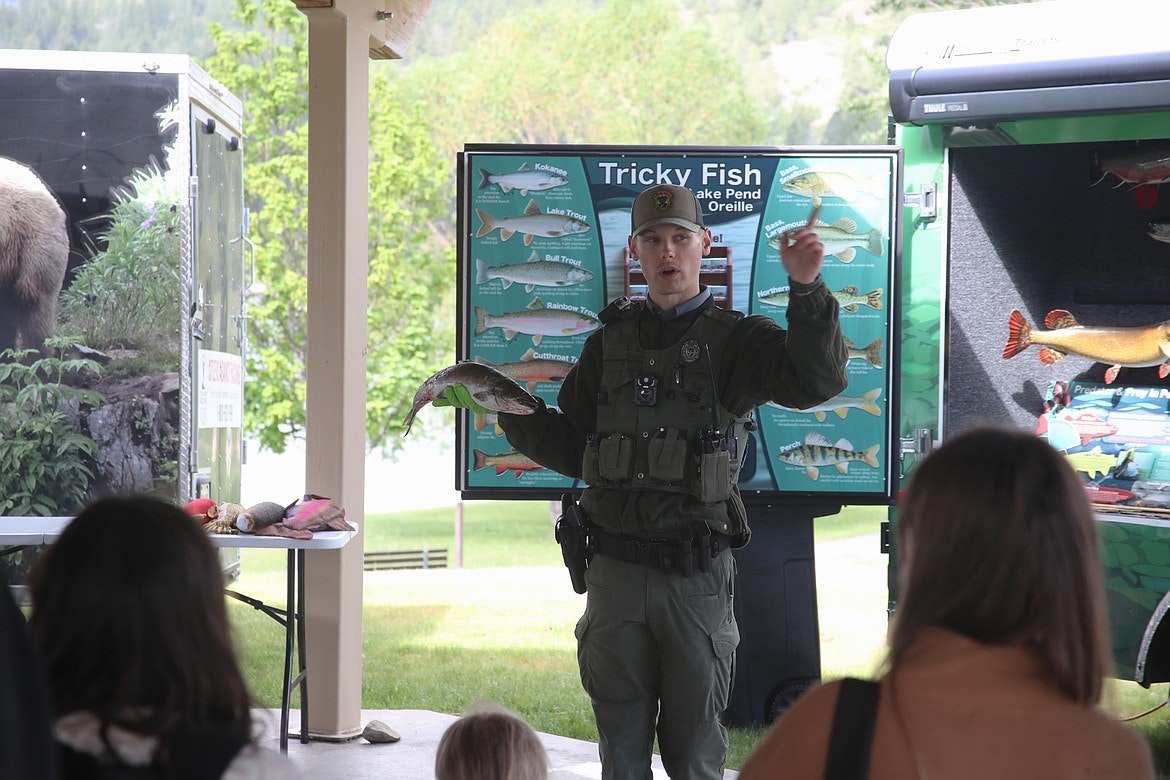 A person in uniform holds a fish, speaking to a group with "Tricky Fish" educational display in the background.