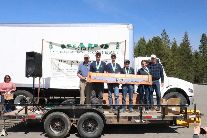 A group of people on a trailer at the Idaho State Forestry Contest with a large saw and medals.