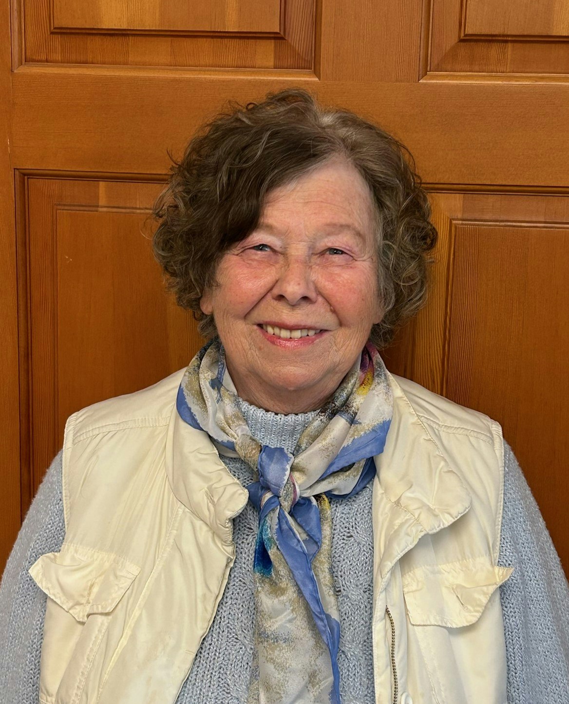 A smiling elderly woman with a scarf and vest, standing in front of a wooden door.