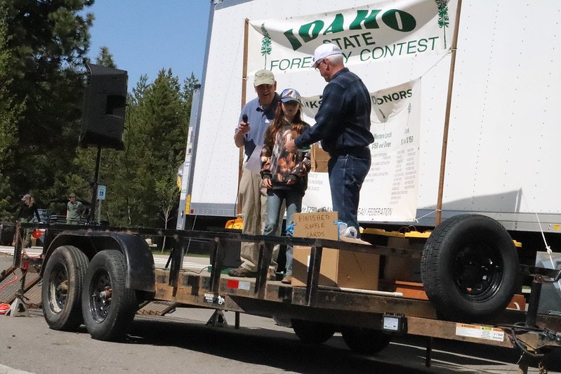 People on a trailer stage at the Idaho State Forestry Contest, with a speaker and a sign.