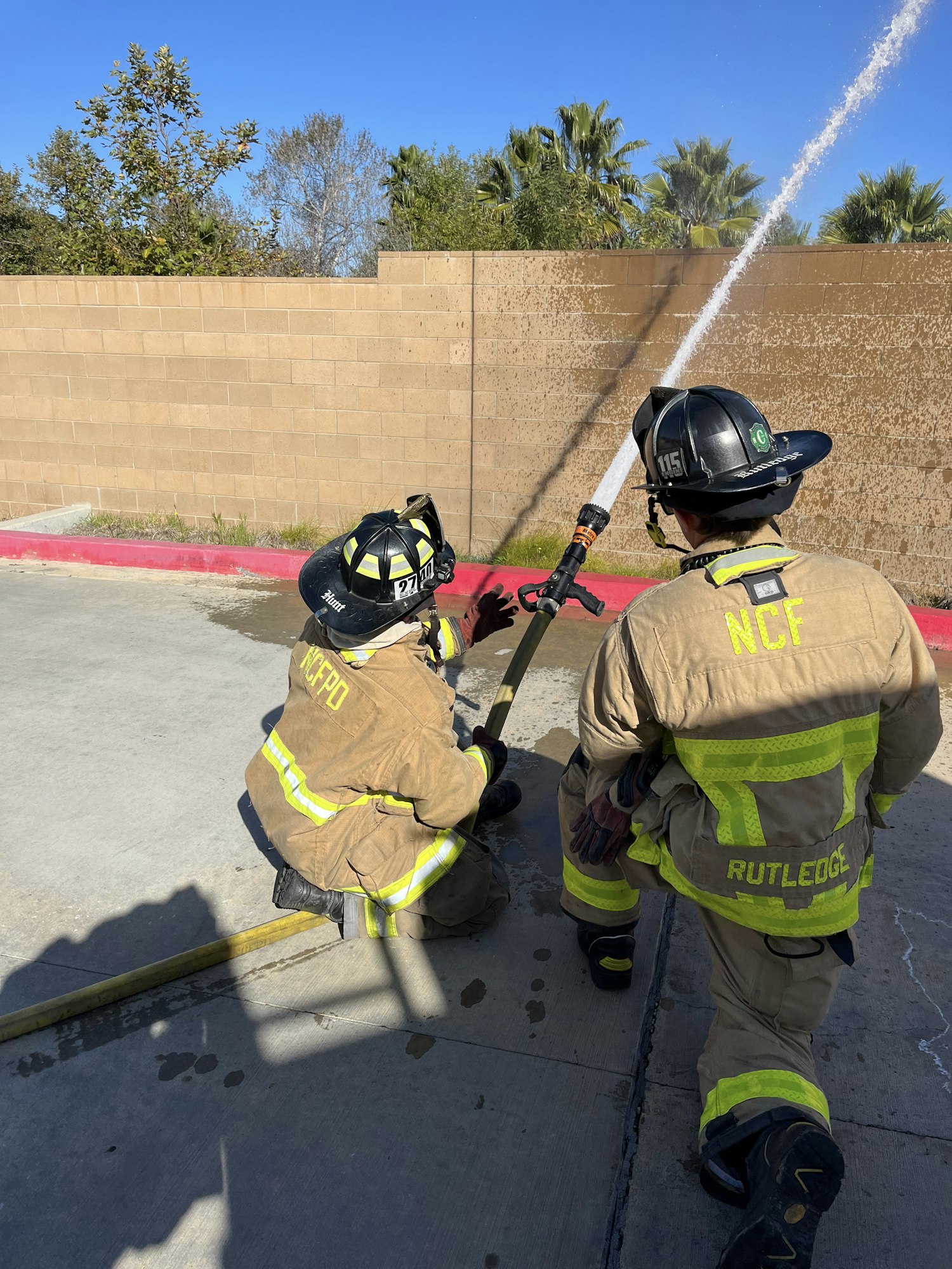 firefighters spraying a fire hose over a wall while kneeling on asphalt