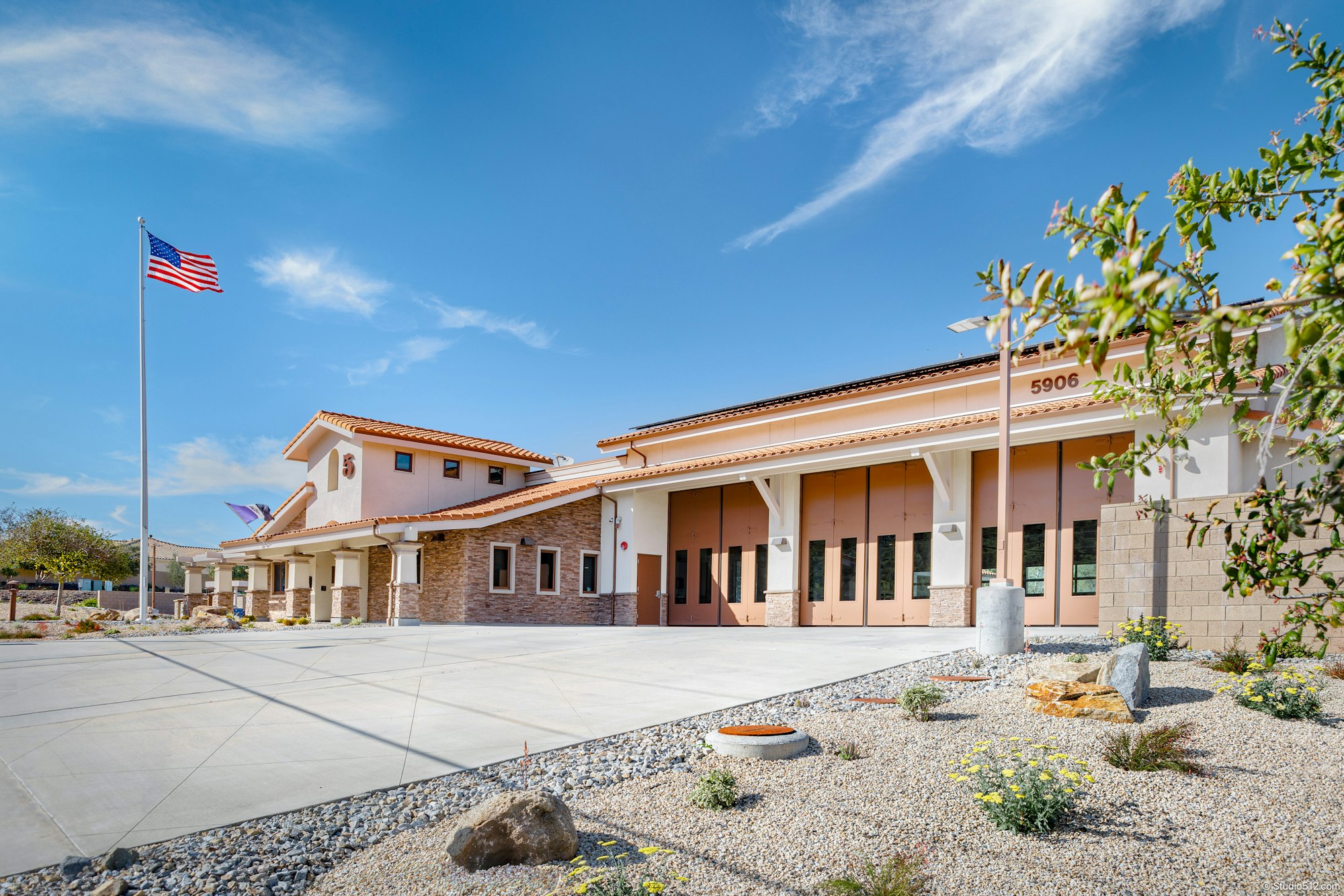 The image shows a modern building with a flag, likely a fire station, featuring a landscaped area and clear blue skies.