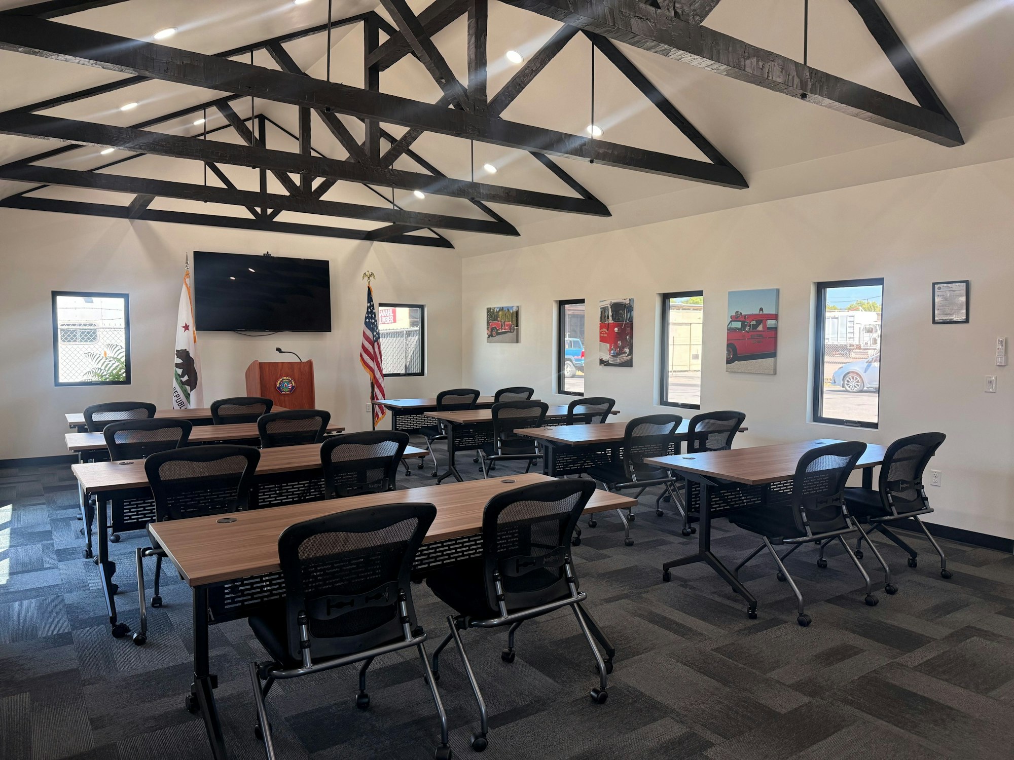 A modern meeting room with tables, chairs, American flags, and wall art, featuring exposed wooden beams and a large screen.