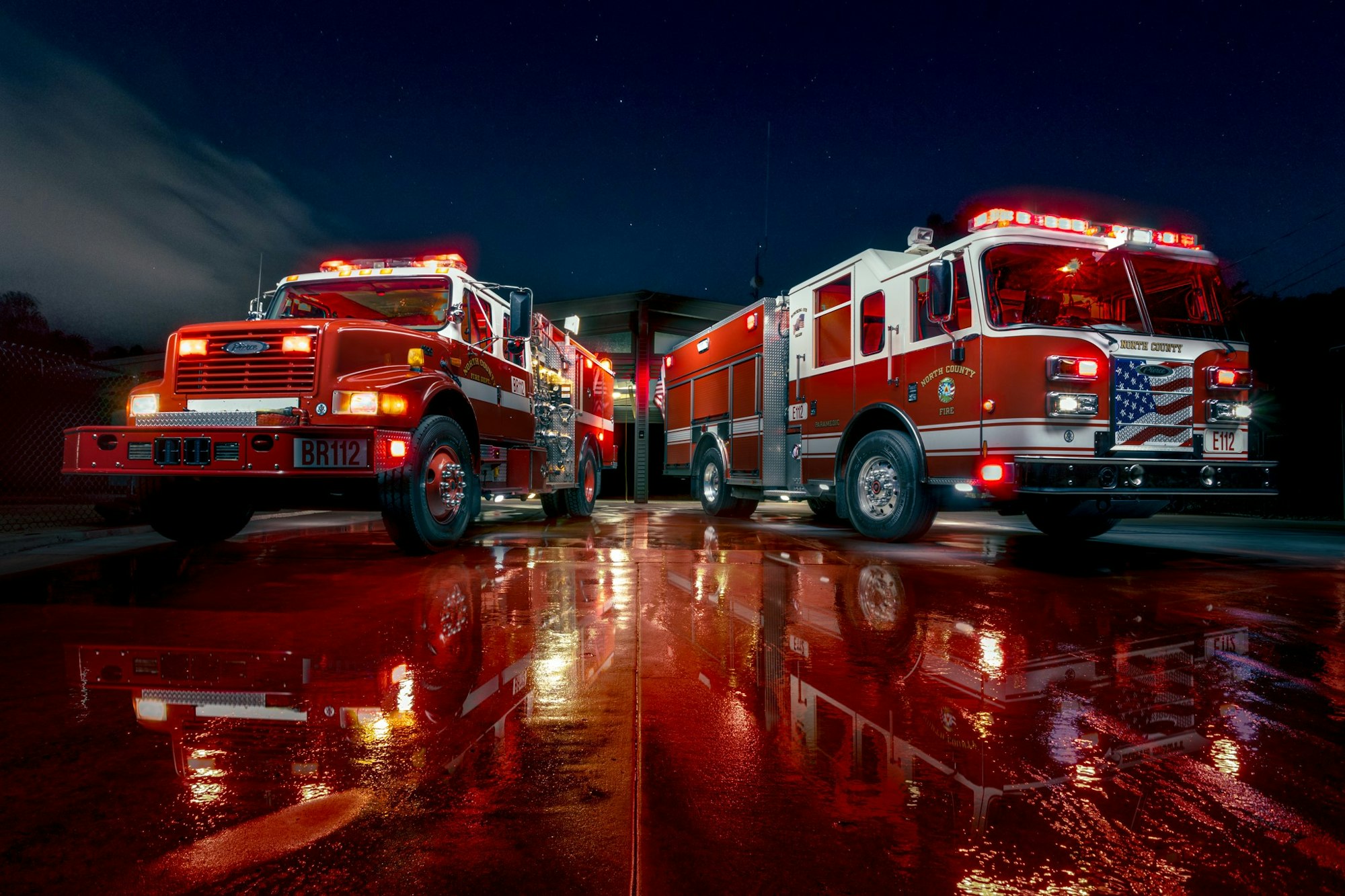 Two red fire trucks on a wet surface with lights on and night sky in the background.