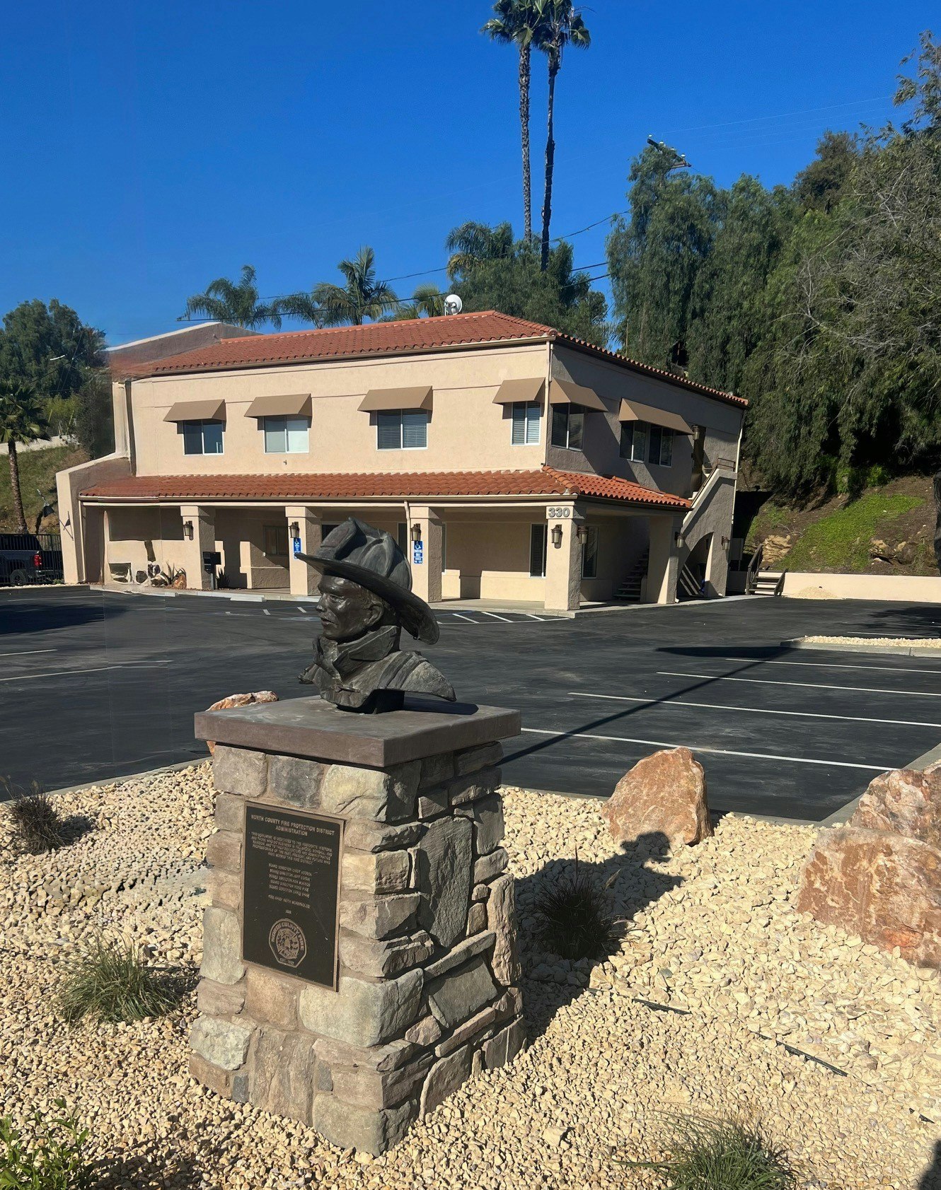 The image features a bust sculpture on a stone pedestal, set in a landscaped area with a building and palm trees in the background.
