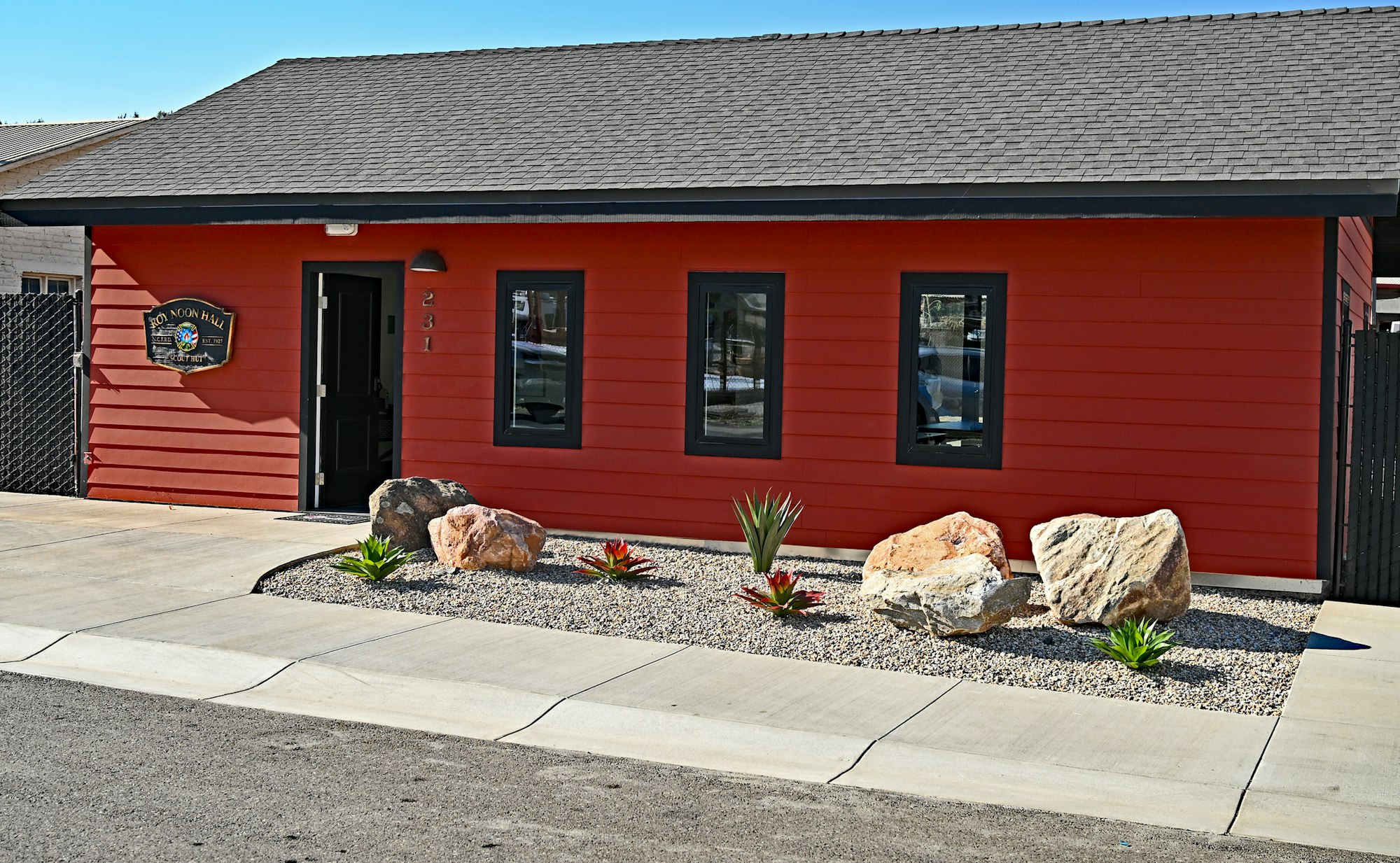 The image shows a red building with a sign, featuring landscaped rocks and plants in front, set against a clear blue sky.