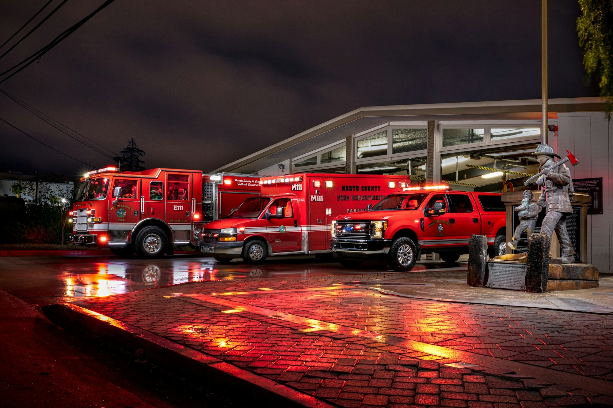 A fire station with emergency vehicles and a firefighter statue at night, wet ground reflecting lights.