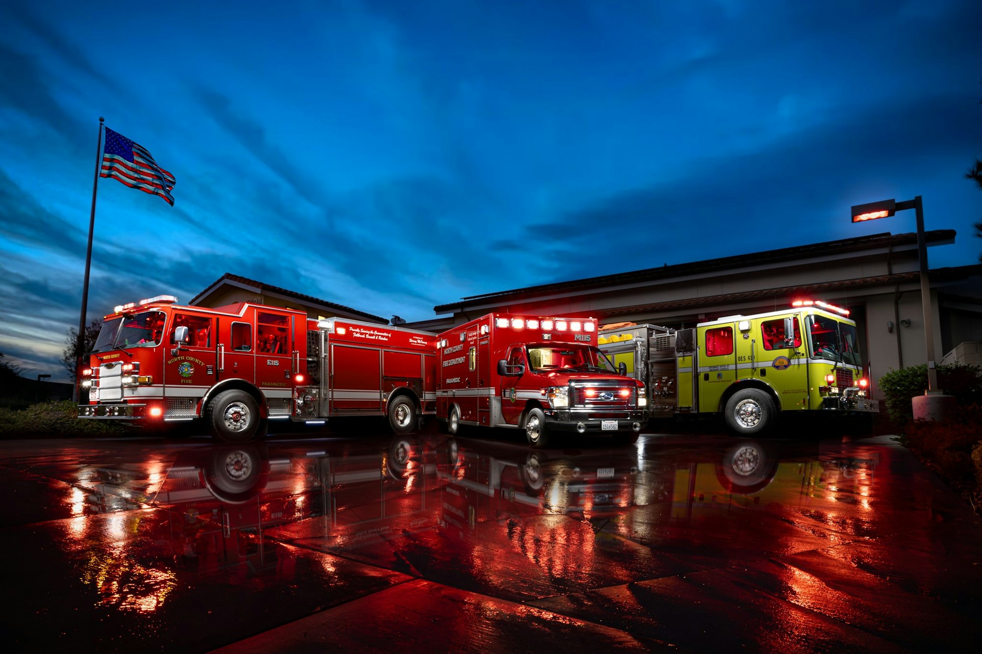 Two fire trucks parked outside a fire station at dusk, wet ground reflecting lights, American flag waving.