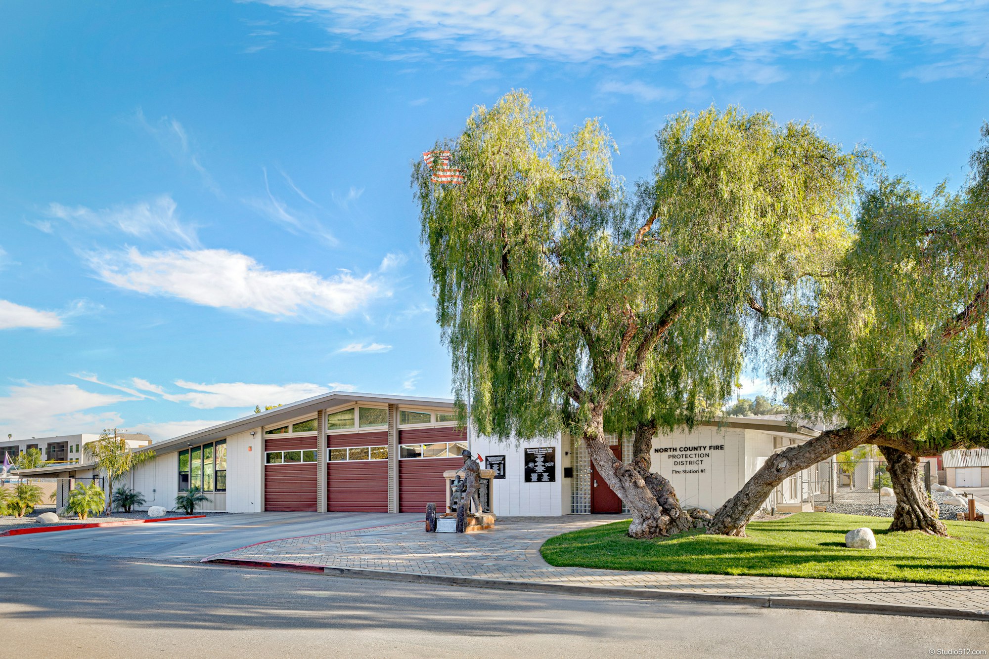 A modern fire station building with a tall tree, an American flag, and a unique sculpture in front, set against a clear blue sky.