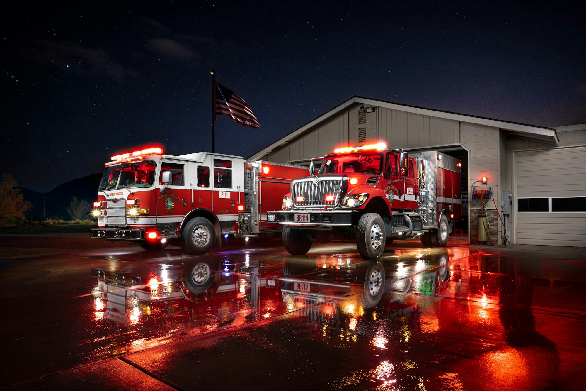 Two fire trucks outside a fire station at night with lights on and wet ground reflecting the scene.