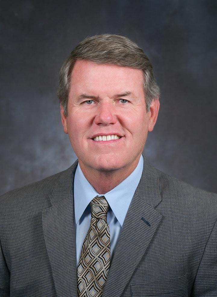 A man in a suit with a patterned tie, smiling at the camera against a dark background.