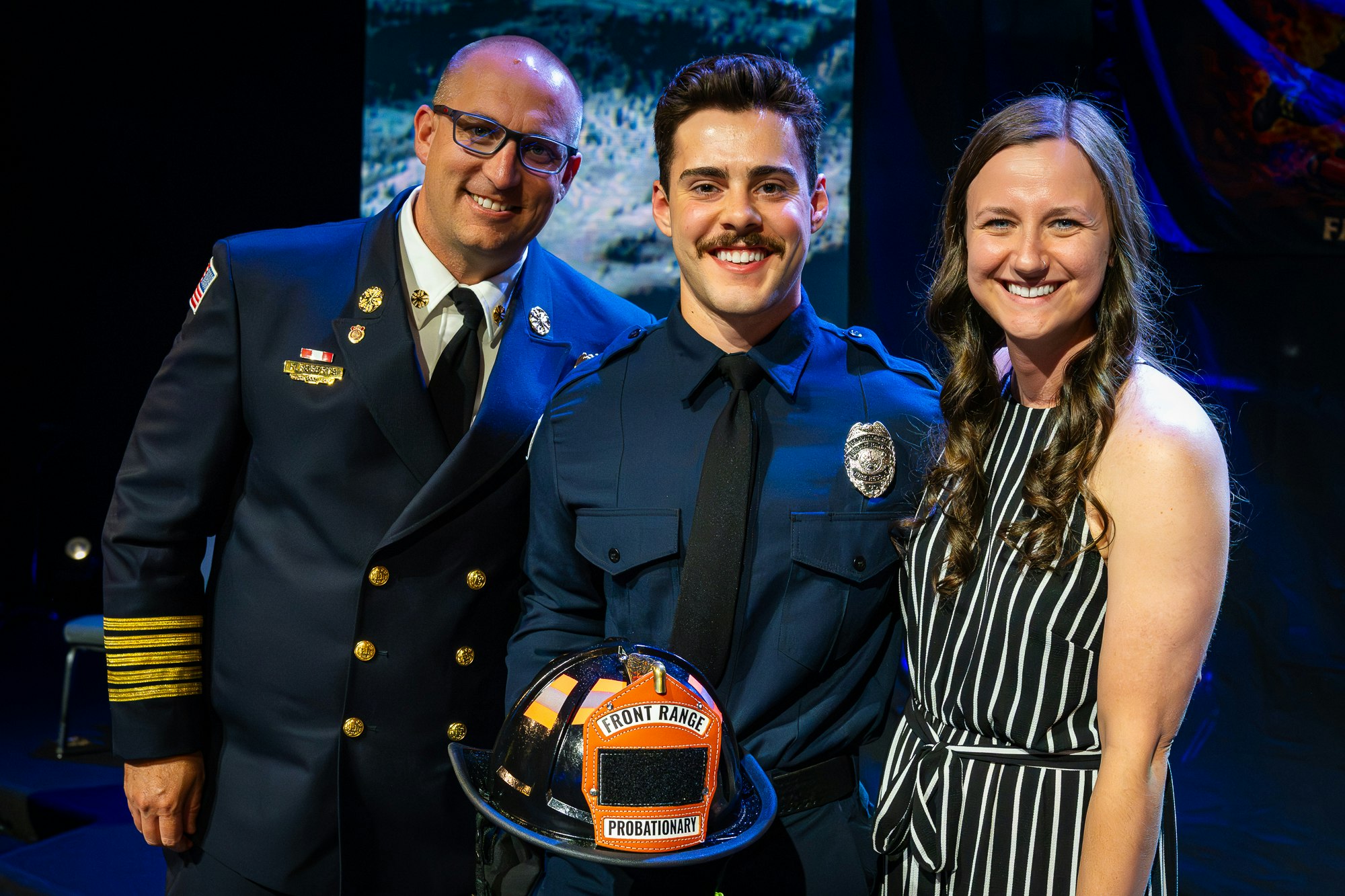 A group photo featuring a fire chief, a new probationary firefighter with a helmet, and a woman smiling beside them.