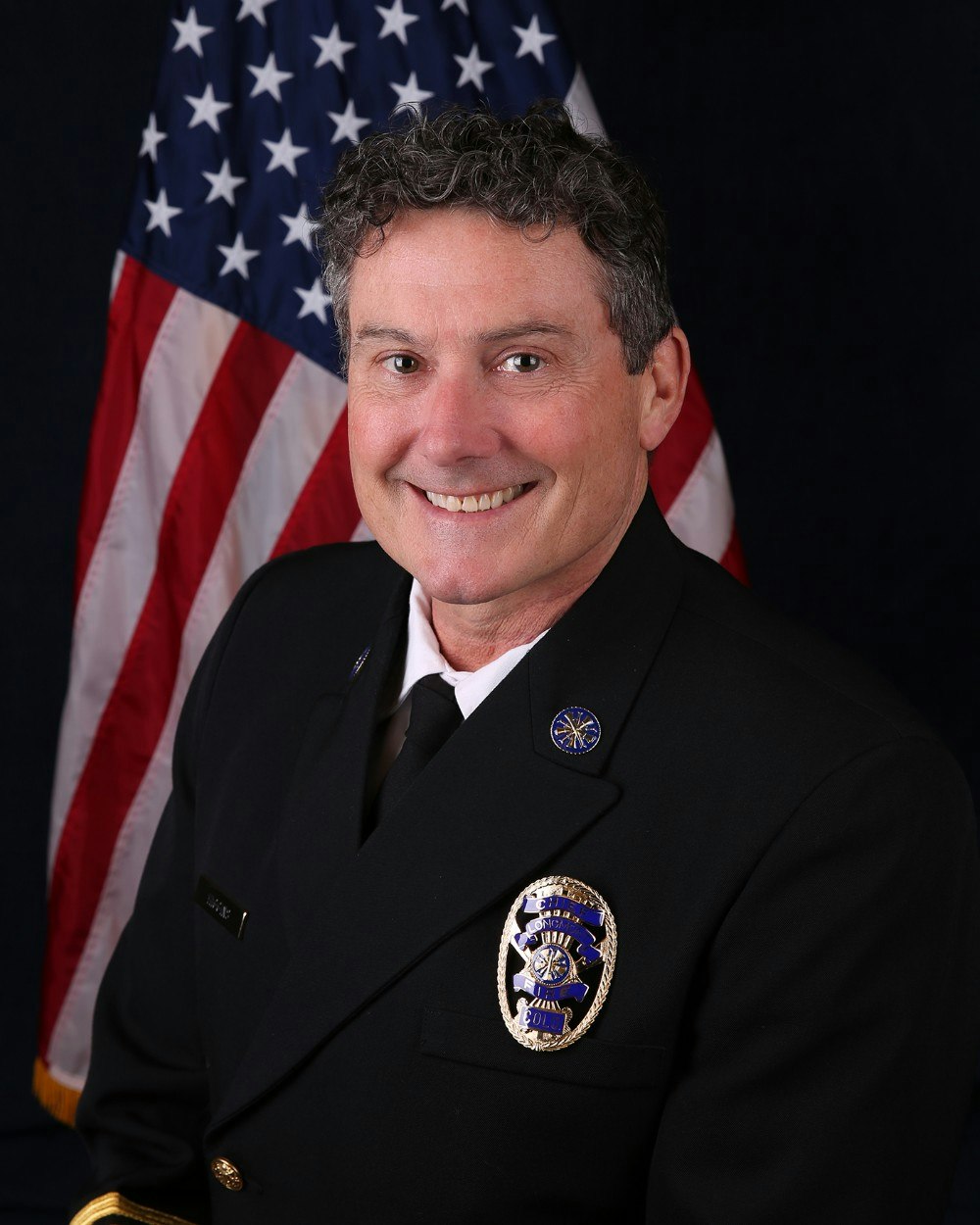 A person in a formal uniform poses smiling in front of an American flag.