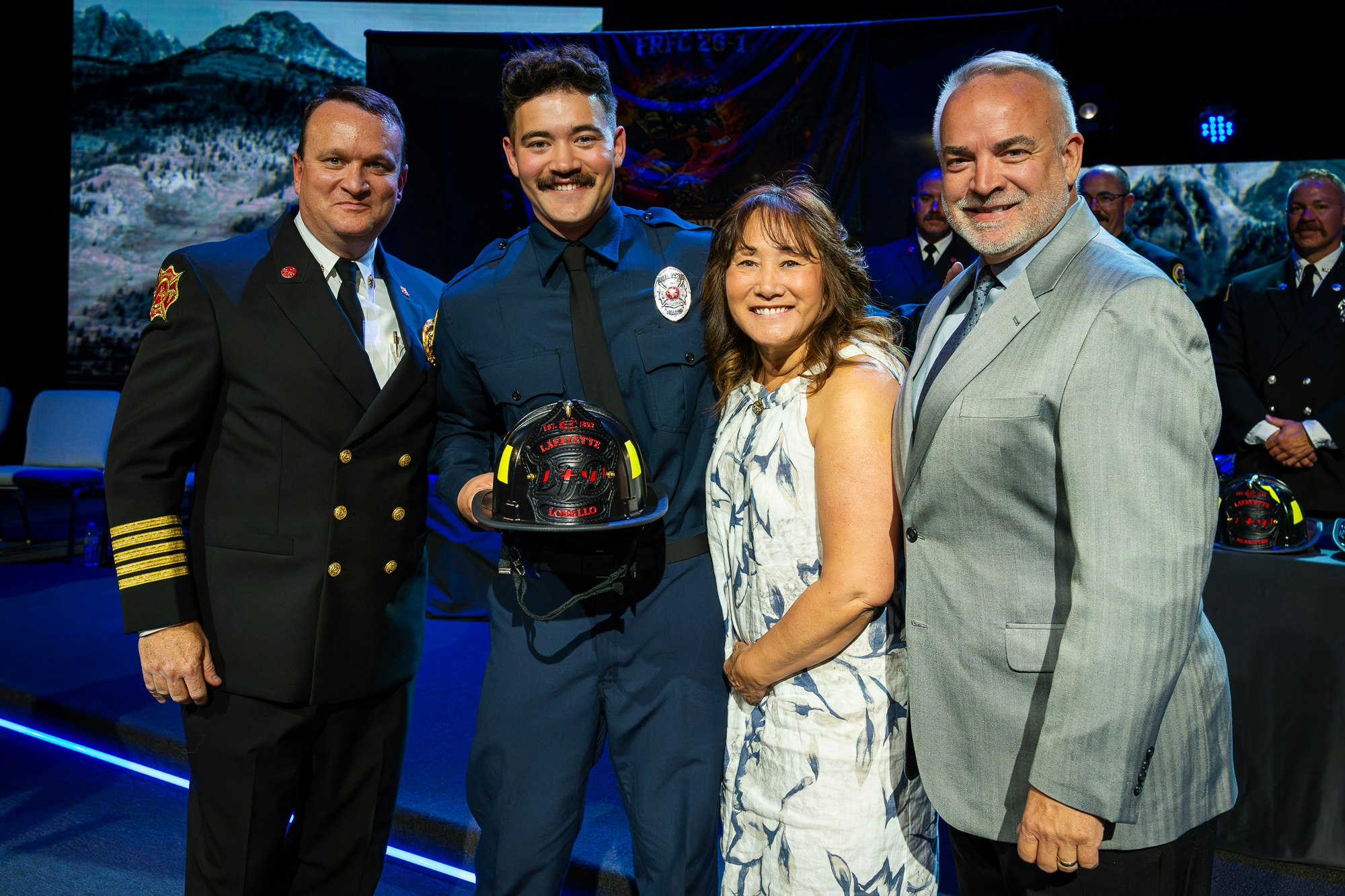 A firefighter receives a helmet at a ceremony, surrounded by family and officials, celebrating a significant achievement.