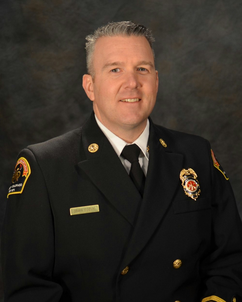A person in a formal uniform with badges and a name tag, posing against a dark background.