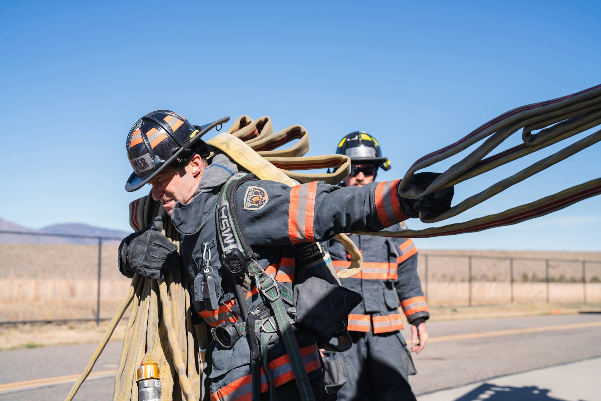 Firefighters in gear carrying hoses on a sunny day.