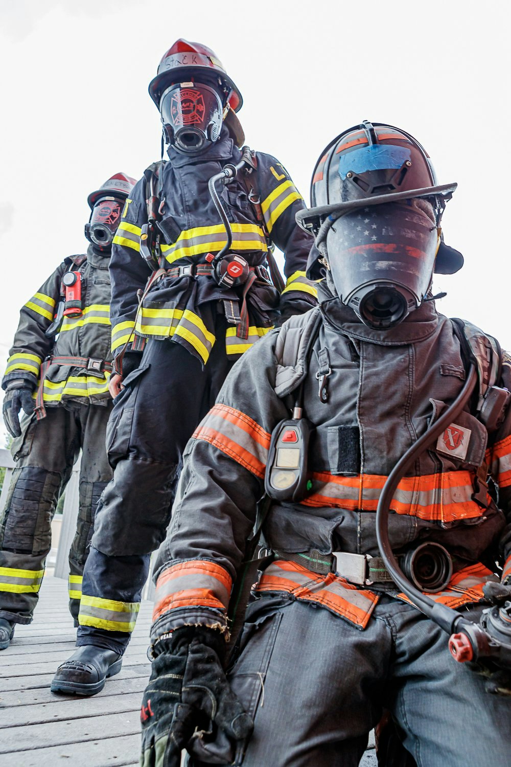 Three firefighters in full gear and face masks stand ready, one kneeling in the foreground.