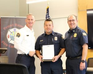 Three people in uniform stand together; the person in the middle is holding a plaque. There is a flag and a banner in the background.